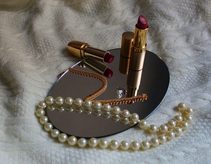 A string of peals, a gold necklace, a pearl earring and two red lipsticks arranged on an upturned mirror on a white bed.