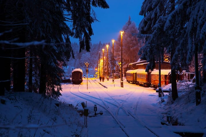 Winter scene: A small train station at dusk illuminated by tall golden lamp posts surrounded by pine trees.