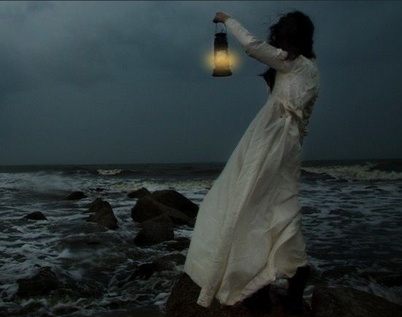 Dark-haired woman in a long white dress, standing on rocks and holding a lantern toward a dark sea.